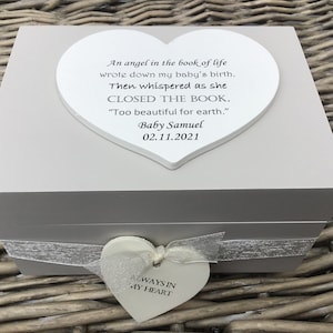 Puede incluir: Una caja de recuerdos gris claro con una tapa blanca en forma de corazón. La tapa tiene una inscripción sobre el nacimiento y el fallecimiento de un bebé. Un corazón más pequeño con "ALWAYS IN MY HEART" cuelga de la caja con una cinta.