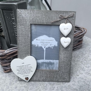 May include: A gray wooden photo frame with a 4 x 6 inch opening, adorned with two heart-shaped ornaments labeled "Elsie" and "Nanna." A larger heart-shaped plaque with a poem is attached. The frame is made from sustainably sourced wood.