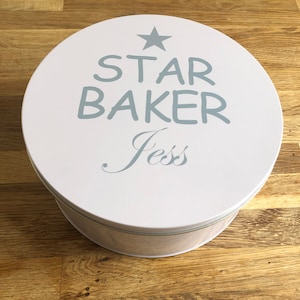 Peut inclure: Une boîte à biscuits ronde en métal blanc avec un couvercle. Le couvercle porte l'inscription "STAR BAKER Jess" en bleu clair, ainsi qu'une étoile. Le bord de la boîte est bleu clair et argenté.