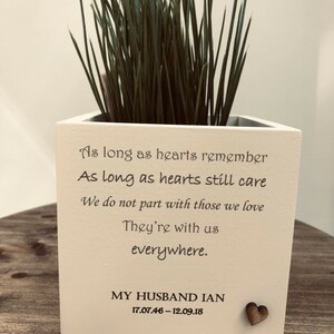 May include: A cream-coloured square planter box with the text "As long as hearts remember...They're with us everywhere." The planter contains green grass. The bottom of the box reads "MY HUSBAND IAN 17.07.46-12.09.18" with a small wooden heart.
