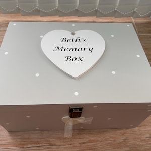 May include: A pale grey memory box with white polka dots and a heart-shaped plaque reading "Beth's Memory Box." The box features a metal clasp and a ribbon tie. A decorative storage item.