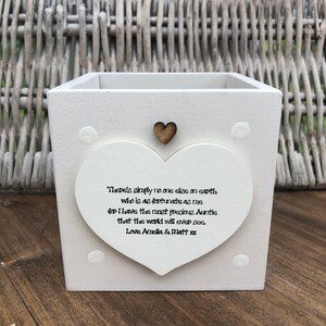 Puede incluir: Una maceta cuadrada de madera blanca con un adorno en forma de corazón. El corazón tiene el texto "There's simply no one else on earth...Love Amelia & Matt xx". Un pequeño corazón de madera está encima del texto. Puntos decorativos en la maceta.