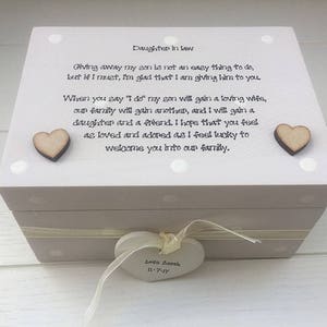 Puede incluir: Una caja de madera gris claro con tapa, decorada con lunares blancos y dos corazones de madera. La caja tiene una cinta y una etiqueta en forma de corazón con texto. La tapa tiene el texto "Daughter in law".