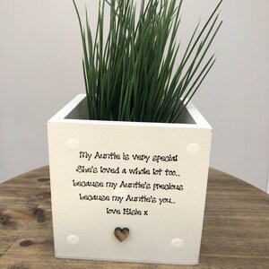 Puede incluir: Una maceta cuadrada blanca con hierba verde, con el texto "My Auntie is very special...love Elsie x." La maceta tiene un pequeño corazón de madera y detalles circulares blancos. La maceta está sobre una superficie de madera.