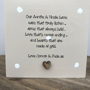 Puede incluir: Placa cuadrada de madera beige con texto en negro: "Our Auntie & Uncle have ears that truly listen..." y un pequeño corazón de madera. Puntos blancos decoran las esquinas.