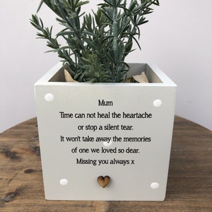May include: A light grey square planter with a green plant. The planter is inscribed with the word "Mum" and a touching message in black text. A small wooden heart is attached, and white dots are on each corner.