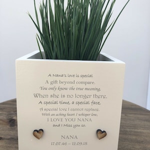 Puede incluir: Una maceta cuadrada de color crema con hierba verde. La maceta tiene un poema y las palabras "I LOVE YOU NANA", el nombre "NANA" y fechas. Dos corazones de madera están adheridos al frente.