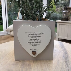 Puede incluir: Una jardinera cuadrada de color gris claro con una placa blanca en forma de corazón. La placa tiene un poema y las palabras "Love Claire x" impresas. La jardinera contiene una planta verde con hojas largas y finas. Un pequeño corazón de madera está adherido a la placa.