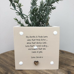 Puede incluir: Una maceta cuadrada beige con una planta verde. La maceta tiene puntos blancos y un corazón de madera. El texto en la maceta dice: "My Auntie & Uncle have ears that truly listen...Love Connor x."