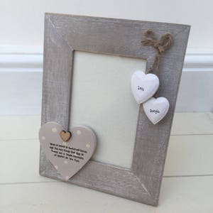Puede incluir: Un marco de fotos de madera gris con adornos en forma de corazón. El marco tiene un acabado gris claro y presenta dos adornos blancos en forma de corazón suspendidos de un lazo de cuerda. Un corazón dice "Love" y el otro dice "Georgia".