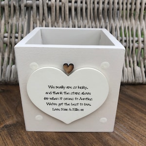 Puede incluir: Una maceta cuadrada de madera blanca con un adorno en forma de corazón. El corazón tiene un pequeño corazón de madera y el texto: "We really are so lucky..." Un artículo decorativo para el hogar.