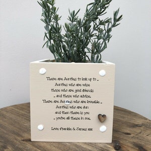 Puede incluir: Una maceta cuadrada beige con una planta verde. La maceta tiene puntos blancos y un corazón de madera. El texto en la maceta dice: "Hay tías... todas sois una." Firmado "Love Frankie & James xx."