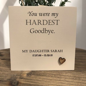 May include: A square, off-white wooden planter box with the text "You were my HARDEST Goodbye." and "MY DAUGHTER SARAH 17.07.66 - 12.09.18." A small wooden heart is affixed to the box. Greenery is planted inside.
