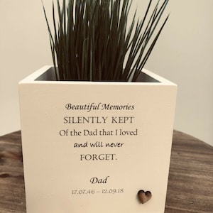 May include: A square, off-white wooden planter box with a plant and the text "Beautiful Memories SILENTLY KEPT Of the Dad that I loved and will never FORGET." Includes the name "Dad" and dates 17.07.46 - 12.09.18. A small wooden heart is affixed to the front.