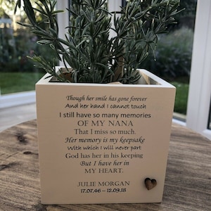 Può includere: Un vaso quadrato bianco sporco con un piccolo cuore di legno, contenente una pianta verde con foglie sottili. Il vaso presenta il testo: "Though her smile has gone forever...OF MY NANA...JULIE MORGAN 17.07.46-12.09.18."