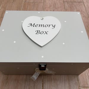 May include: A light gray memory box with a white heart-shaped plaque that reads "Memory Box." The box has a polka dot pattern and a silver clasp with a ribbon. A decorative storage box for keepsakes.
