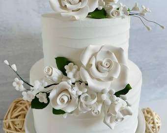 Puede incluir: Pastel blanco de dos pisos decorado con grandes rosas blancas de azúcar, pequeñas flores blancas y hojas verdes. El pastel tiene un diseño texturizado y acanalado y se asienta sobre un soporte blanco para pasteles. Un postre decorativo.