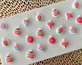 May include: Assortment of 18 Christmas ornament-shaped decorations. The ornaments are white with red, green, and blue designs. The ornaments are arranged on a white surface.