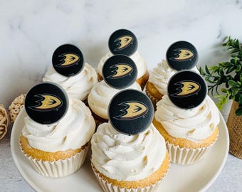 Op de afbeelding: Acht cupcakes met witte frosting staan op een wit bord. Elke cupcake is versierd met een zwart en goud logo. De cupcakes zitten in gele papieren vormpjes. Een kleine groene plant staat op de achtergrond.