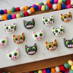 Puede incluir: Una colecci&oacute;n de 11 botones con forma de gato de colores con varios patrones y colores, incluyendo blanco, negro, marr&oacute;n y verde. Los botones est&aacute;n dispuestos sobre una superficie blanca.