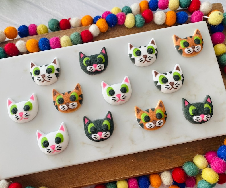 Puede incluir: Una colecci&oacute;n de 11 botones con forma de gato de colores con varios patrones y colores, incluyendo blanco, negro, marr&oacute;n y verde. Los botones est&aacute;n dispuestos sobre una superficie blanca.