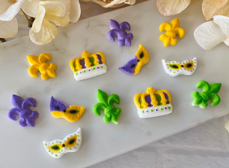 May include: Purple, green, and yellow sugar decorations shaped like fleur-de-lis, crowns, and masks. The decorations are on a white surface.