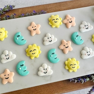 Può includere: Una collezione di 15 biscotti di zucchero color pastello decorati con facce sorridenti. I biscotti hanno la forma di stelle, lune, nuvole e soli.