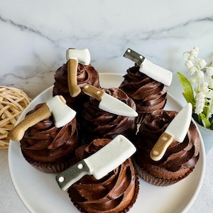 May include: Six chocolate cupcakes decorated with white fondant knives and axes. The cupcakes are arranged on a white plate.