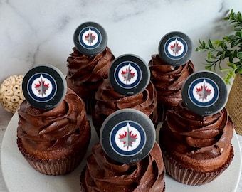 Op de afbeelding: Zes chocolade cupcakes met swirl frosting, elk bekroond met een hockey puck-vormige decoratie met het Winnipeg Jets logo. De cupcakes zijn gerangschikt op een witte plaat, perfect voor een hockey themafeest.