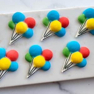 Pode incluir: Seis biscoitos em forma de balão decorados com glacê vibrante azul, amarelo, verde e vermelho. Cada biscoito apresenta uma base triangular branca com detalhes de linhas pretas, representando fios de balão, dispostos sobre uma superfície branca.