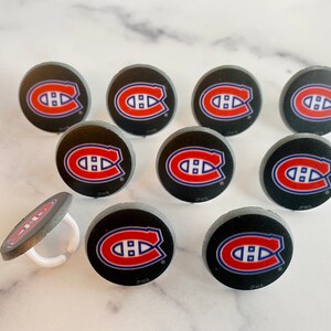 Op de afbeelding: Tien ronde zwarte en grijze hockeypuck cupcake toppers met een rood, wit en blauw Montreal Canadiens logo. E&eacute;n topper heeft een witte plastic ring.