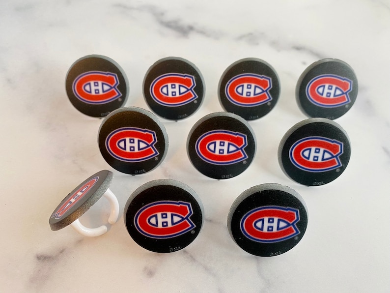 Op de afbeelding: Tien ronde zwarte en grijze hockeypuck cupcake toppers met een rood, wit en blauw Montreal Canadiens logo. E&eacute;n topper heeft een witte plastic ring.