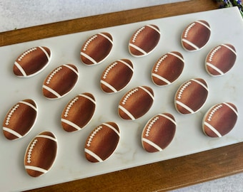 May include: A collection of fifteen oval-shaped cookies decorated to resemble brown American footballs. Each cookie features a textured brown surface with white icing details representing the laces and stripes at both ends of the football.