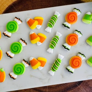 May include: A selection of Halloween-themed felt decorations, including orange pumpkins, green and white swirl sweets, candy corn, green and white striped sweets, and orange swirl sweets. The decorations are arranged on a white surface.
