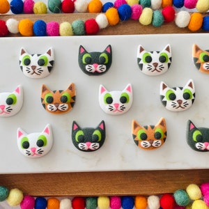 Puede incluir: Doce galletas de az&uacute;car con forma de cara de gato de colores sobre una superficie blanca. Las galletas est&aacute;n decoradas con glaseado blanco, negro, verde y naranja.