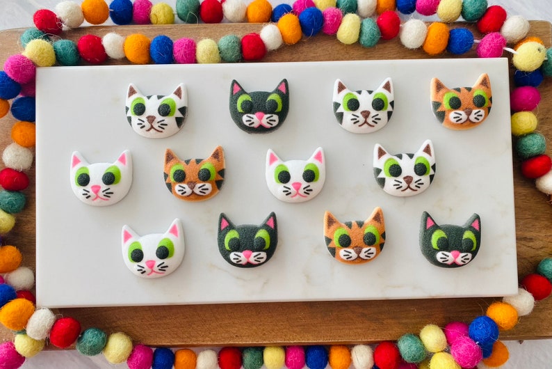 Puede incluir: Doce galletas de az&uacute;car con forma de cara de gato de colores sobre una superficie blanca. Las galletas est&aacute;n decoradas con glaseado blanco, negro, verde y naranja.