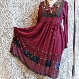 May include: A burgundy long-sleeve dress with a V-neck and embroidered neckline. The dress features a gathered waist and a patchwork hemline with floral and geometric patterns. The dress is displayed on a mannequin.