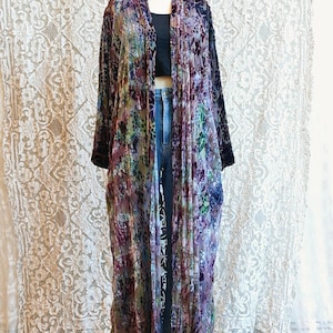 May include: A long, open-front kimono-style robe with a floral pattern in shades of purple, green, and blue. The robe has long sleeves and a velvet texture. The garment is displayed on a mannequin, paired with a black top and jeans.