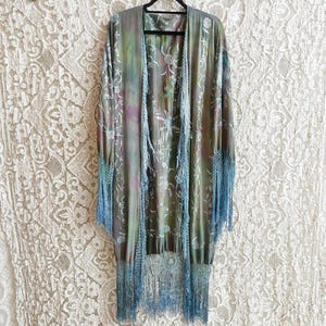 May include: A long, flowing kimono-style garment with a tie-dye effect in shades of green, purple, and brown. The kimono features intricate floral embroidery and is trimmed with long, light blue fringe. It hangs against a white lace backdrop.