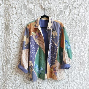Puede incluir: Una camisa de manga larga con botones de varios colores con un diseño de patchwork. La camisa presenta pájaros, mariposas y patrones geométricos bordados en tonos azules, verdes, naranjas y marrones.
