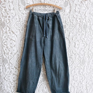 May include: Dark teal wide-leg pants with a drawstring waist and two front pockets. The pants are hanging on a wooden hanger against a white lace background. The pants have a relaxed fit and a casual style.