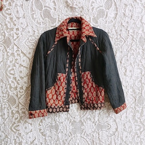 Puede incluir: Chaqueta acolchada negra con detalles florales rojos y marrones. La chaqueta tiene cuello y cierre de botones. El interior y los bolsillos muestran el diseño floral. La chaqueta está colgada en una percha negra.