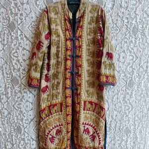 Puede incluir: Un abrigo largo beige con un estampado de elefantes repetido en rojo y amarillo. El abrigo tiene cuello mandarín, ribete azul oscuro y cierres de rana en la parte delantera. El abrigo está colgado de una percha negra.