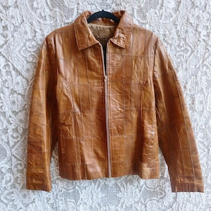 May include: A tan leather jacket constructed from a patchwork of rectangular panels. The jacket features a classic fold-down collar, long sleeves, and a full-length vertical zipper closure down the center front. It has two side pockets.