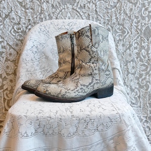 Peut inclure: Paire de bottines à motif peau de serpent, dans des tons gris et beige. Les bottines ont une fermeture éclair latérale et un petit talon. Elles sont posées sur une chaise recouverte de dentelle blanche.