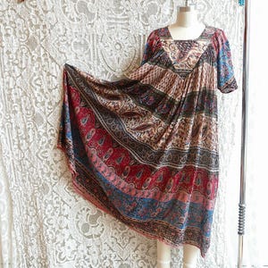 Könnte beinhalten: Vintage, gemustertes Kleid in Rot-, Blau- und Beigetönen, präsentiert auf einer Schaufensterpuppe. Das Kleid hat einen quadratischen Ausschnitt und kurze Ärmel. Der Stoff hat ein Paisley- und Blumenmuster. Das Kleid hängt an einer Metall-Kleiderstange.