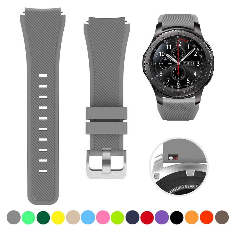 May include: A grey silicone watch strap with a diagonal ribbed texture, featuring a silver-toned metal buckle and a quick-release pin mechanism. The strap is shown attached to a black smartwatch with a round face, alongside a color palette of fifteen circular swatches in various solid colors including grey, green, yellow, blue, pink, purple, black, orange, and brown.