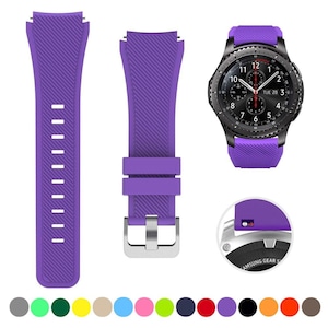 May include: A purple silicone watch band with a diagonal ribbed texture, featuring a silver-toned metal buckle and a quick-release pin mechanism. The band is shown attached to a black smartwatch with a round analog-style face displaying the date.