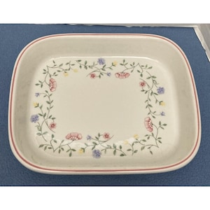May include: Rectangular ceramic baking dish with a floral design. The dish has a cream-colored interior and a red rim. The floral pattern includes pink, blue, and yellow flowers with green vines, creating a decorative border.