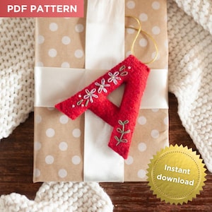 May include: A red felt letter A ornament featuring white and green floral embroidery and a gold hanging cord. The letter is placed on a brown gift box with white polka dots and a white ribbon, labeled with PDF PATTERN text.
